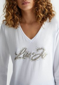 White long-sleeve shirt with a v-neck, featuring a decorative design of metallic studs forming cursive text in the center. Soft texture.