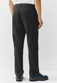 Man met zwarte rechte broek en donkerblauwe leren schoenen, van achteren weergegeven tegen een effen witte achtergrond.
