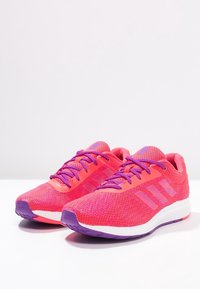 Baskets de sport roses avec une tige en maille texturée, accents violets, logo à trois bandes et une semelle intermédiaire blanche avec une semelle extérieure violette.