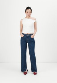 Femme debout les mains dans les poches, portant un haut blanc à une épaule, un jean bleu foncé avec des fentes à l'avant, et des escarpins rouges sur fond blanc.