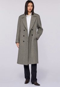 Szary płaszcz dwurzędowy z mieszanki wełny, z szerokimi klapami, sześcioma czarnymi guzikami i bocznymi kieszeniami; model stoi z rękami w kieszeniach, w zestawieniu z ciemnymi spodniami.