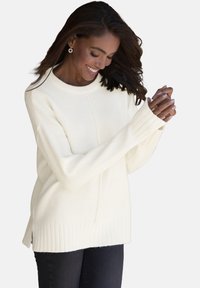 Cremfarbener Strickpullover mit Rundhalsausschnitt, geripptem Saum, seitlichen Schlitzen und langen Ärmeln, getragen über dunklen Jeans.