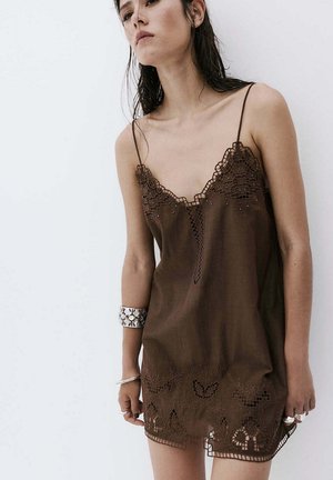 Giovane donna con capelli bagnati che indossa un vestito marrone senza maniche con dettagli in pizzo e braccialetti d’argento al polso sinistro.