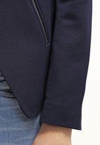 Námořnický blejzr s texturovanou tkaninou, který obsahuje detail se zipem na rukávu a klasické řemeslné přizpůsobení. Viditelný šev a design manžet.