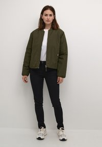 Oliiviroheline nahkjakk, millel on tõmblukk, kantud valge t-särgi ja mustade kitsaste teksade peal, koos valgete ja hallide spordijalatsitega.