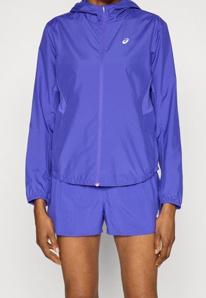 Person trägt eine violette Kapuzenjacke mit Reißverschluss und passende Shorts mit weißen Markenlogos auf der Brust und am linken Oberschenkel.