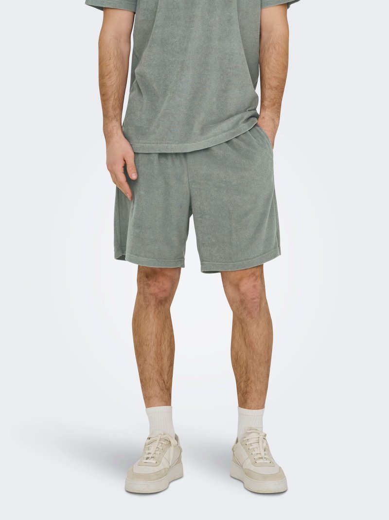 Homme portant un short en éponge gris et une chemise à manches courtes assortie, des chaussettes blanches et des baskets beiges, debout avec une main dans la poche.