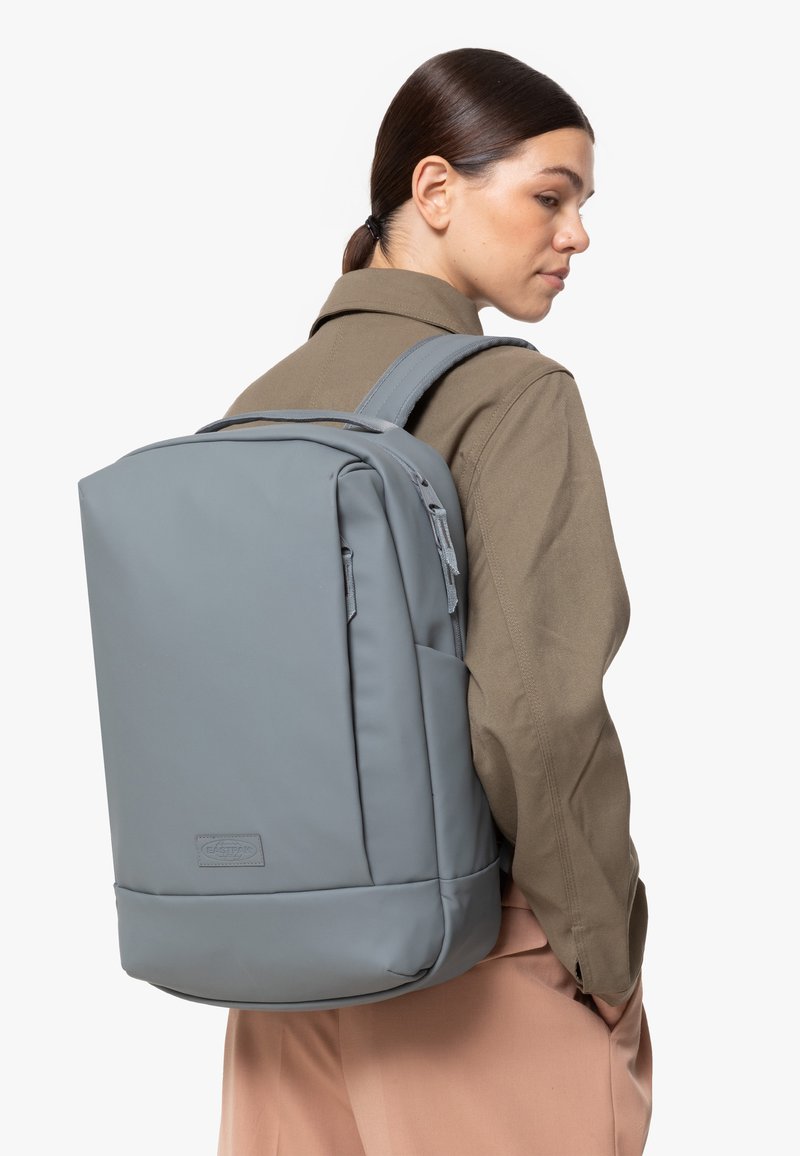 Šedý batoh vyrobený z hladkého, odolného materiálu s obdélníkovým tvarem, nastavitelnými ramenními popruhy a přední kapsou na zip.