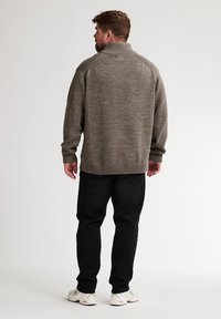 Uomo con barba che indossa un maglione di maglia marrone, pantaloni neri e sneakers bianche, in piedi con le spalle rivolte verso un fondo chiaro e semplice.