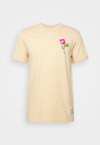 Beżowa bawełniana koszulka z różowym kwadratowym logo i zielonym wzorem rośliny po lewej stronie klatki piersiowej. Krótkie rękawy i okrągły dekolt.