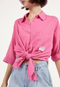 Kobieta w różowej koszuli zapinanej na guziki, związanej z przodu w węzeł, z podwiniętymi rękawami oraz jasnoniebieskich spodniach z wysokim stanem.