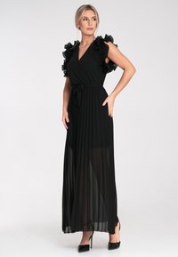 Schwarzes, plissiertes Maxikleid mit Rüschenärmeln, Wickeldesign und gebundenem Taillenband. Transparente Textur, fließender Rock und hochhackige Schuhe gut sichtbar.