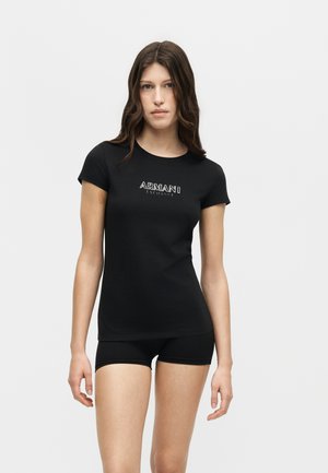 Jeune femme aux longs cheveux bruns portant un t-shirt noir ajusté Armani Exchange et un short noir sur un fond gris uni.