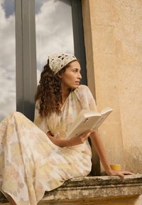Femme en robe fleurie et bandeau en crochet assise près de la fenêtre, lisant un livre, avec une tasse jaune à côté d'elle sur le rebord en pierre.