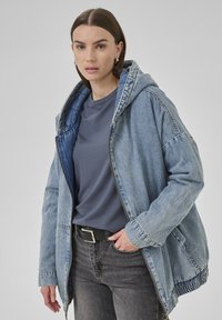 Giacca di denim con cappuccio, colore azzurro chiaro, presenta una chiusura con zip, orlo elasticizzato e maniche ampie, abbinata a una t-shirt grigia e jeans neri.