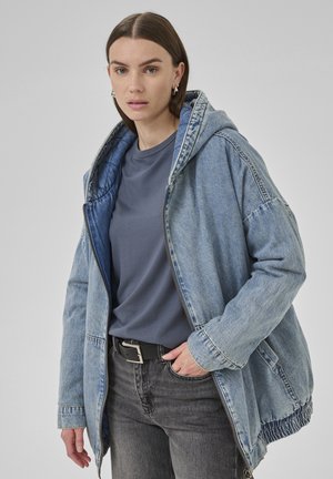 Denim jacket with a hood, light blue color, features a zip closure, elasticized hem, and roomy sleeves, paired with a gray t-shirt and black jeans.