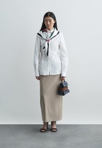 Λευκό πουκάμισο με ρυτιδωτή υφή, μπεζ μολυβιού φούστα, μαύρες σανδάλια, κρατώντας μια μικρή μαύρη τσάντα με καφέ λεπτομέρειες και μαντίλι γύρω από τον λαιμό.