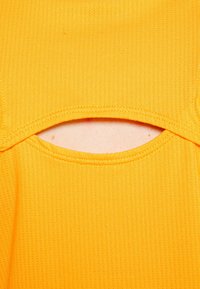 Žlutá žebrovaná tkanina má kulatý výstřih s horizontálním výřezem, který odhaluje malou část kůže pod otvorem.