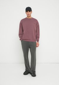 Rotes Sweatshirt mit Rundhalsausschnitt und gerippten Bündchen, kombiniert mit grauen Hosen und schwarzen Sneakers. Das Model steht vor einem weißen Hintergrund.