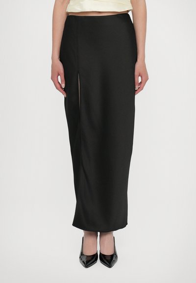 Naine kannab kõrge vöökohaga musta maksiseelikut küljesildusega, millele on lisatud mustad terava varbaga kontsakingad ja ta seisab ühevärvilise tausta ees.