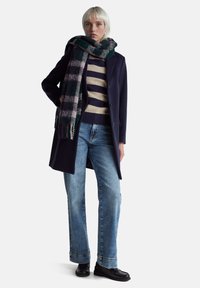 Osoba ubrana w granatowy płaszcz, pasiasty sweter, niebieskie dżinsy, czarne buty i duży zielono-fioletowy kraciasty szalik, stojąca na tle jednolitego tła.