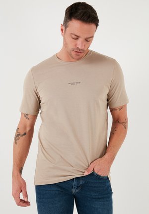 Tricou bej cu maneci scurte, realizat dintr-un material moale, având un guler de tip crew și un mic imprimeu de text negru pe piept. Purtați cu blugi albaștri.