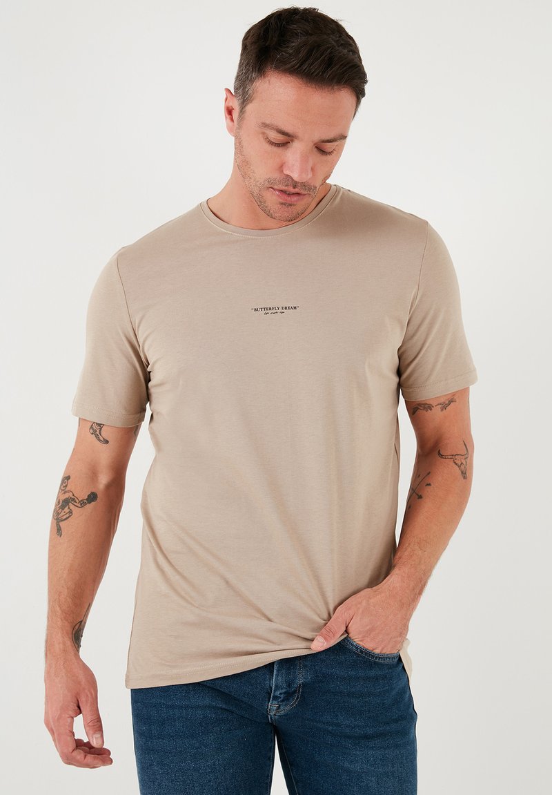 Tricou bej cu maneci scurte, realizat dintr-un material moale, având un guler de tip crew și un mic imprimeu de text negru pe piept. Purtați cu blugi albaștri.