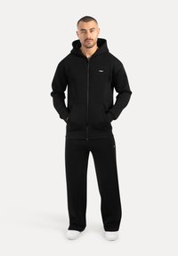 Muž stojící s rukama v kapsách, oblečený v černé mikině na zip, černých širokých kalhotách a bílých teniskách na jednobarevném pozadí.