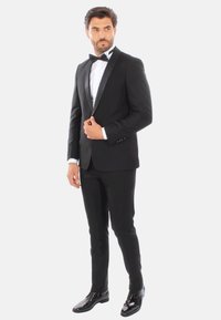 Tuxedo nero con rever in satin, camicia bianca e papillon nero. Pantaloni aderenti e scarpe eleganti nere lucide completano l'outfit.