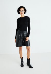 Zwarte langärmbloes en leren shorts, gecombineerd met zwarte enkellaarsjes. De shorts hebben voorzakken en een textuurafwerking.