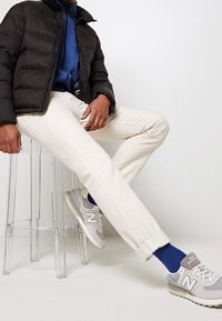 Person som sitter på en genomskinlig pall och bär en svart puffjacka, blå tröja, krämfärgade byxor, blå strumpor och grå New Balance-sneakers.