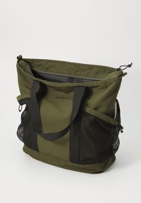 Sac fourre-tout olive avec fermeture éclair, deux poignées en tissu noir, et poches latérales en maille pour le stockage. Matériau durable et léger.