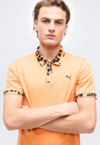 Jeune homme aux bras croisés portant un polo orange avec des bordures florales foncées sur le col et les manches, se tenant devant un fond uni.