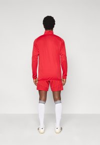 Κόκκινο αθλητικό τοπ φόρμας με ψηλό γιακά, συνδυασμένο με κόκκινα σορτς. Λευκές κάλτσες μέχρι το γόνατο με τρεις μαύρες ρίγες και λευκά ποδοσφαιρικά παπούτσια.