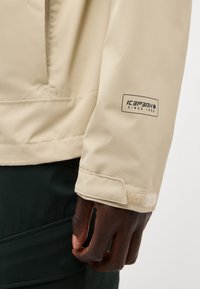 Manche de veste beige avec le logo « ICEPEAK SINCE 1996 », poignet réglable en Velcro, et main à peau foncée reposant à côté d'un pantalon noir.