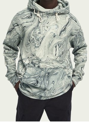 Mężczyzna ubrany w szarą i czarną bluzę z wzorem marmuru, z rękami w kieszeniach i czarne spodnie na jednolitym tle.