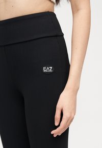 Κοντινό πλάνο ενός ατόμου που φοράει μαύρο ριμπ κολάν με ψηλή μέση και λογότυπο EA7 Emporio Armani στο μηρό.