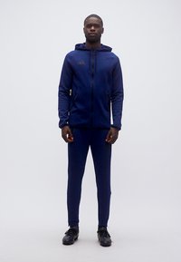 Άνδρας όρθιος, στραμμένος προς τα εμπρός, με μπλε ναυτικό φόρμα Adidas με κουκούλα και μαύρα αθλητικά παπούτσια, σε απλό φωτεινό φόντο.