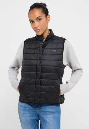 Czarna puchowa kamizelka z wysokim kołnierzem, pikowanym wzorem, przednimi kieszeniami i subtelnym logo po lewej stronie. Noszona na szarym, długim rękawie.