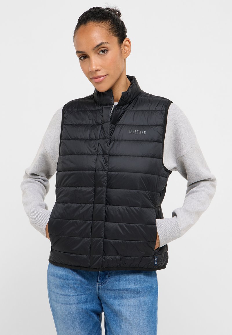 Czarna puchowa kamizelka z wysokim kołnierzem, pikowanym wzorem, przednimi kieszeniami i subtelnym logo po lewej stronie. Noszona na szarym, długim rękawie.