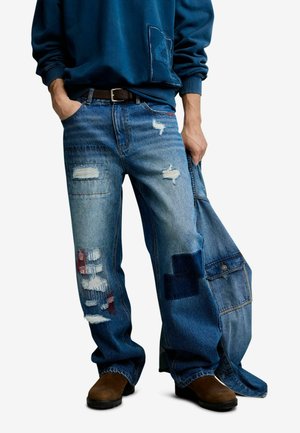 Personne portant un jean bleu usé avec des détails en patchwork, des chaussures marron et un sweat-shirt bleu, tenant une veste en denim dans une main.