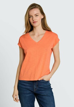 Frau mit langen blonden Haaren, die ein oranges kurzärmeliges V-Ausschnitt-Shirt und blaue Jeans trägt, steht mit einer Hand in der Tasche.