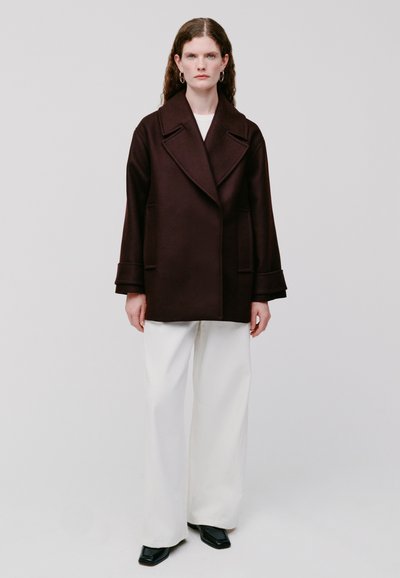 Cappotto in lana marrone con un ampio colletto, taglio oversize e tasche laterali, abbinato a pantaloni bianchi a gamba larga e stivaletti neri con tacco.