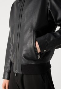 Schwarze Leder-Bomberjacke mit strukturierter Oberfläche, Reißverschlusstaschen und gerippten schwarzen Manschetten. Verfügt über einen Reißverschluss vorne.