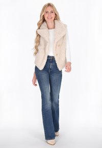 Mujer con cabello largo y rubio, vistiendo un chaleco de sherpa color crema, una camiseta de manga larga blanca, jeans azules y zapatos beige, de pie contra un fondo blanco.