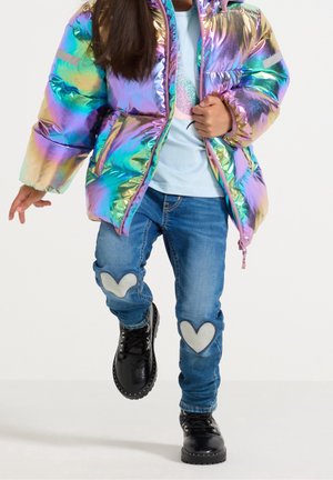 Enfant portant une doudoune irisée, un jean bleu avec des patches cœurs blancs aux genoux et des bottines noires à lacets marchant vers l'avant.
