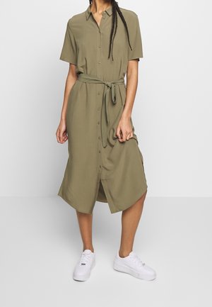 Kobieta nosząca oliwkową, zapinaną na guziki sukienkę midi z krótkimi rękawami i przewiązanym paskiem w talii, w połączeniu z białymi trampkami, stojąca na jednolitym tle.