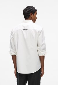 Chemise blanche à manches longues avec manches retroussées, ourlet arrondi et étiquette au dos. Texture en coton lisse, design simple sans motifs.