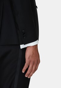 Svart skräddarsydd kostym med en slim-fit jacka och byxor. Noterbara detaljer inkluderar fem svarta knappdetaljer på ärmarna och vita skjortärmar.