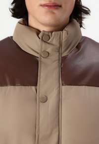 Padded Jacke mit hohem Kragen, featuring einen beigen Körper und braunen glänzenden Schultereinsätzen. Mit metallischen Druckknöpfen verschlossen.
